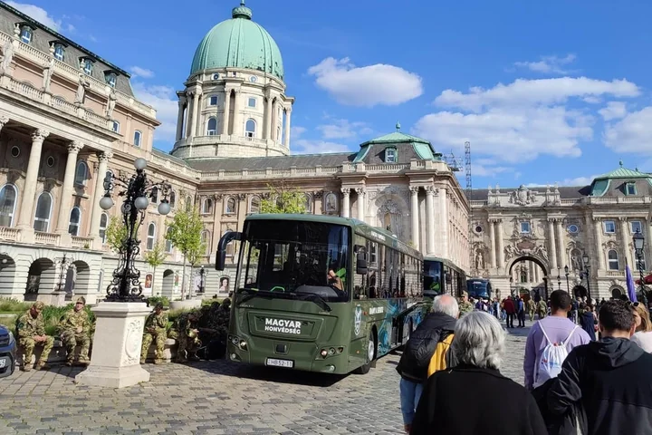 A bus parked in the courtyard of the Royal Palace below the green dome of Buda Castle, with visitors walking past