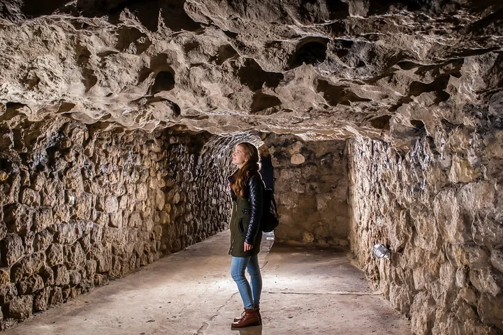 A visitor standing in a narrow lit passage with rough limestone walls inside the Buda Castle Cave